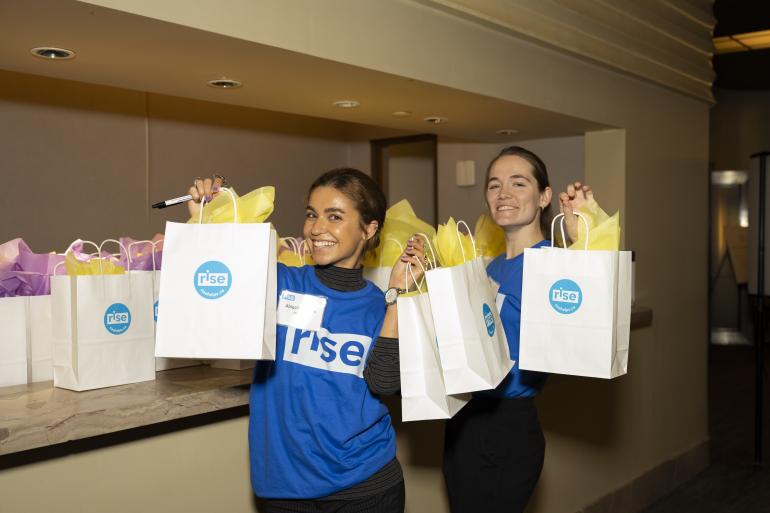 Two women smiling in Rise shirts holding gift bags
