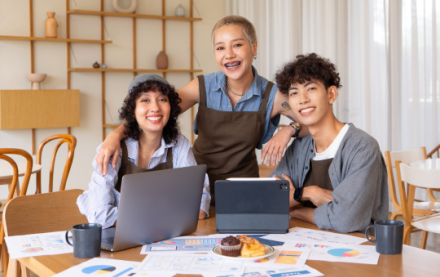 Three young people at a desk covered in papers and devices, working together