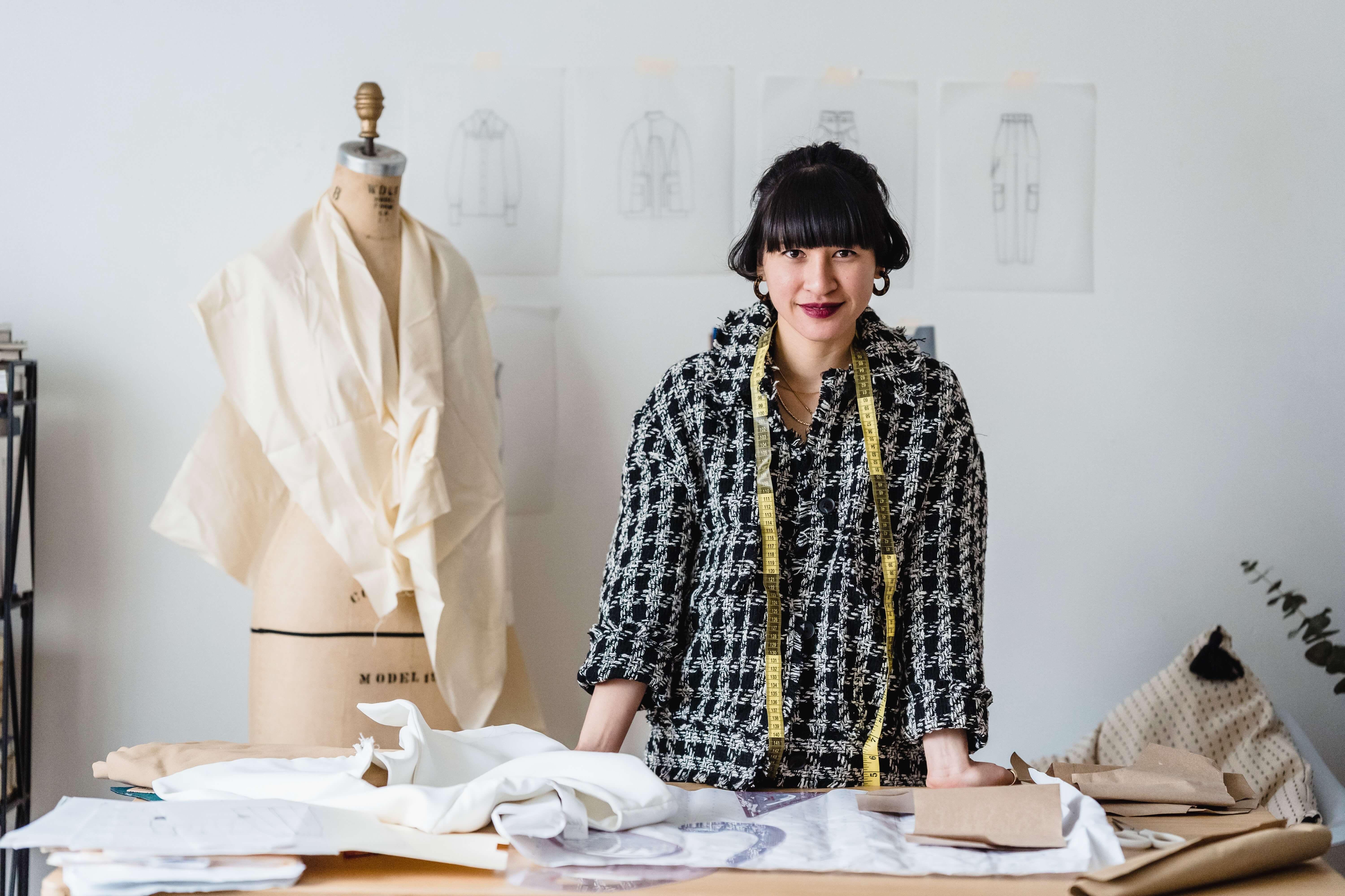 A photo of a woman standing over a sewing table, with a dress form next to her.